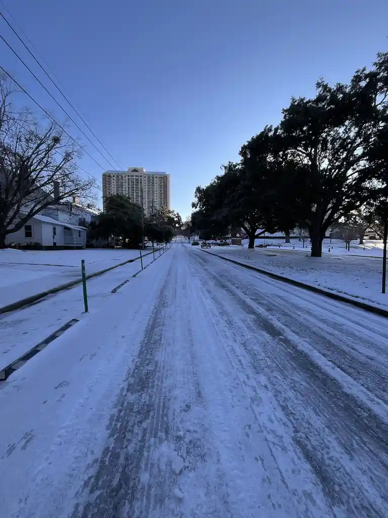 A snow-covered residential street with tire tracks leading toward a distant high-rise building under a clear blue sky…