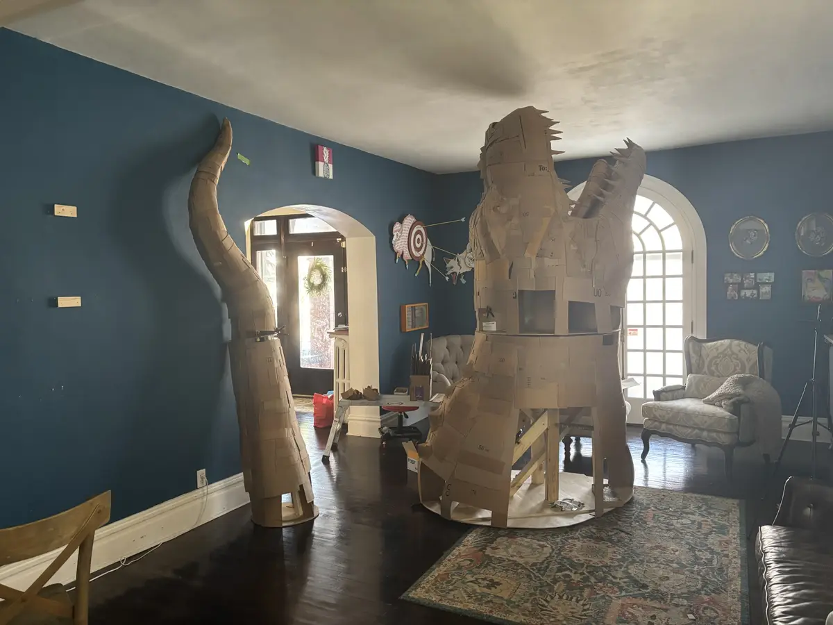 A large, unfinished cardboard sculpture of a dinosaur-like creature stands in a living room, its head and tail visible, with…