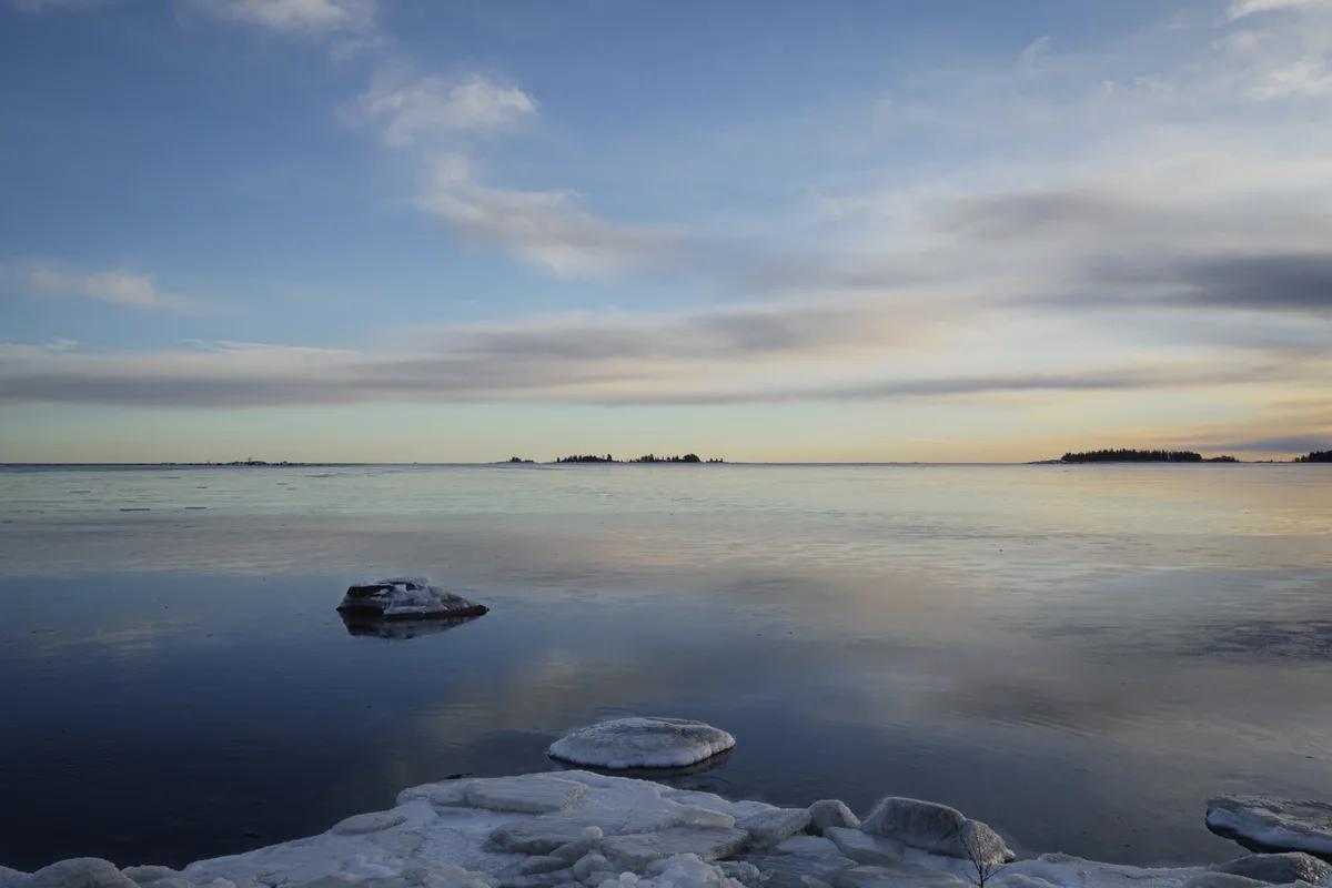 A serene winter landscape shows a frozen lake with ice chunks and snow-covered rocks in the foreground, reflecting a calm…