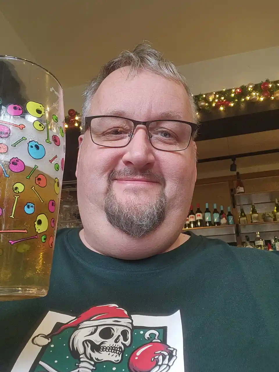 A man with glasses and a goatee smiles while holding a large beer glass decorated with colorful cartoon skulls and bones. He…