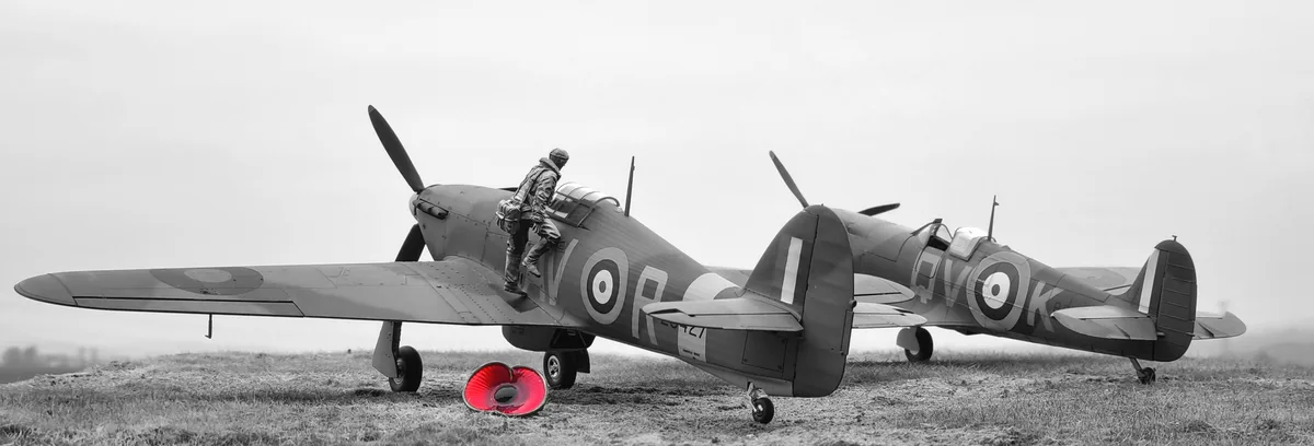 A black-and-white photo shows two vintage British fighter planes, likely Hurricanes, parked on a grassy field. A statue of a…