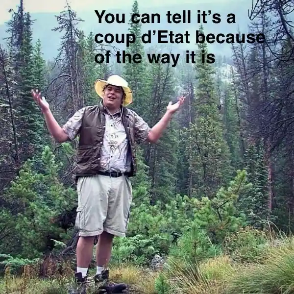 A man in a yellow hat, vest, and shorts stands on a grassy hillside in a forest, arms outstretched in a gesture of dramatic…