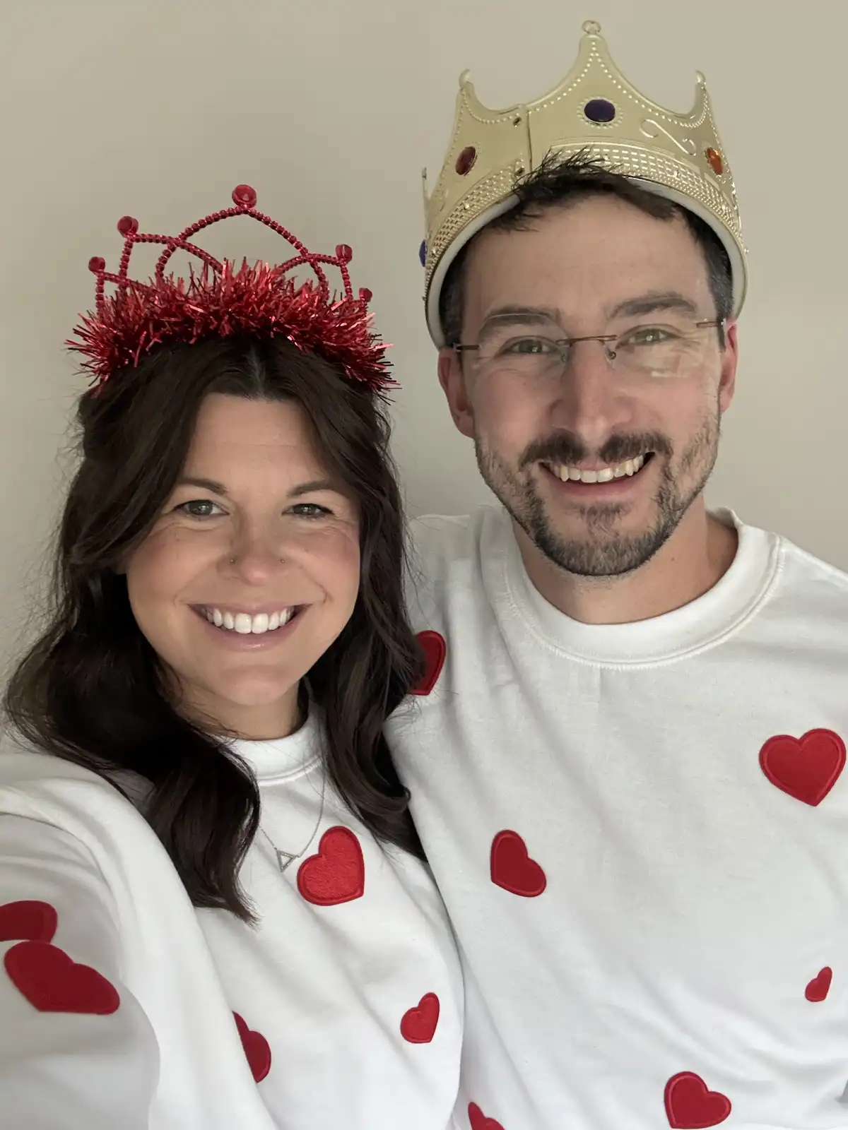 A smiling couple poses for a selfie, both wearing white sweaters adorned with red heart patches. The woman wears a sparkly…