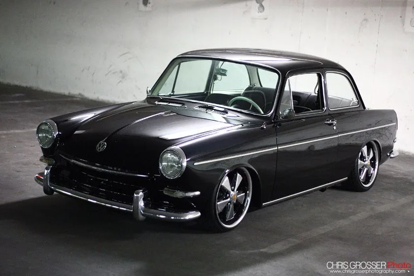 A glossy black classic Volkswagen Beetle with custom chrome wheels and lowered suspension is parked in a dimly lit garage…