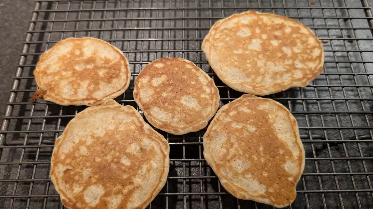 Five golden-brown pancakes rest on a black metal cooling rack, their surfaces showing uneven browning and slightly irregular…