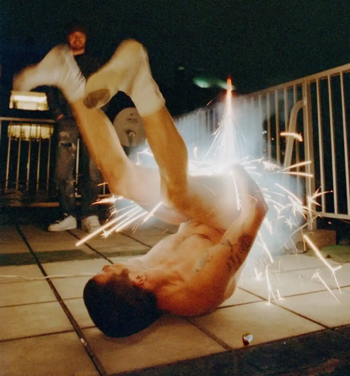 A shirtless man is captured mid-flip on a tiled floor at night, with bright sparks flying around him as if from a firework…