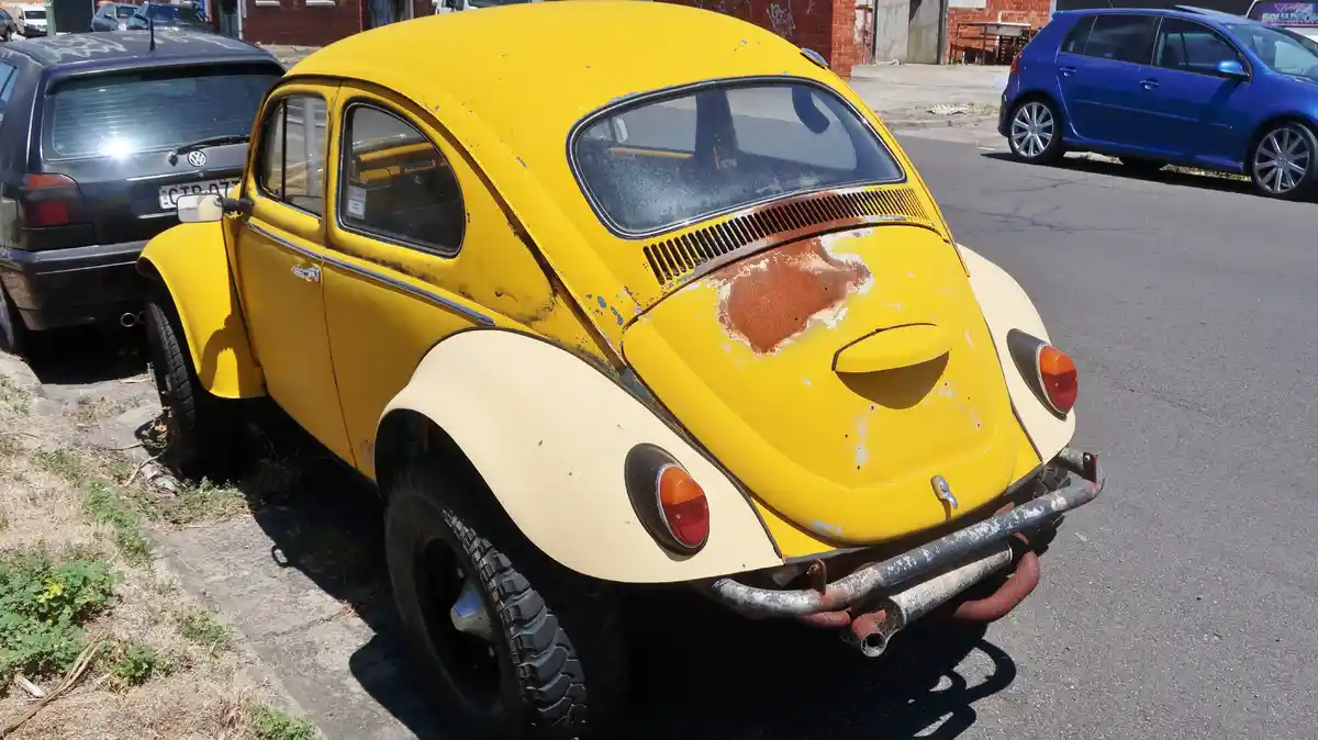 A yellow and cream vintage Volkswagen Beetle with visible rust and wear is parked on a street, featuring rugged off-road…