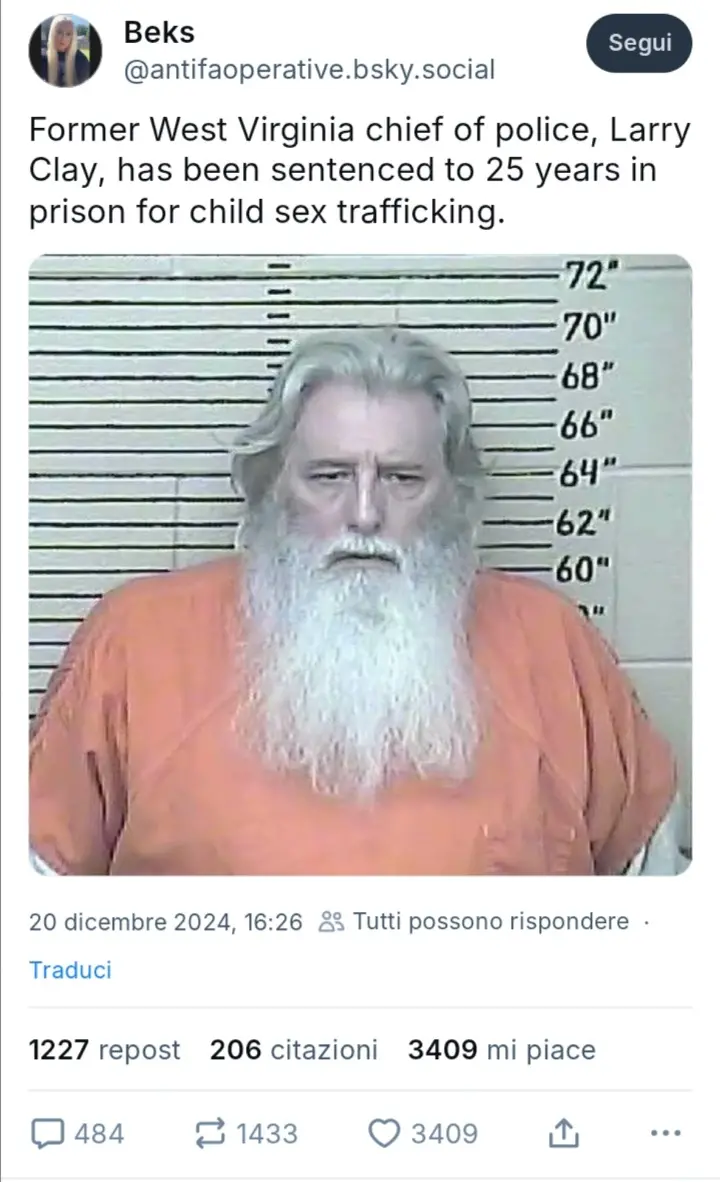 A mugshot of a man with long white hair and a full white beard, wearing an orange prison jumpsuit, standing in front of a…