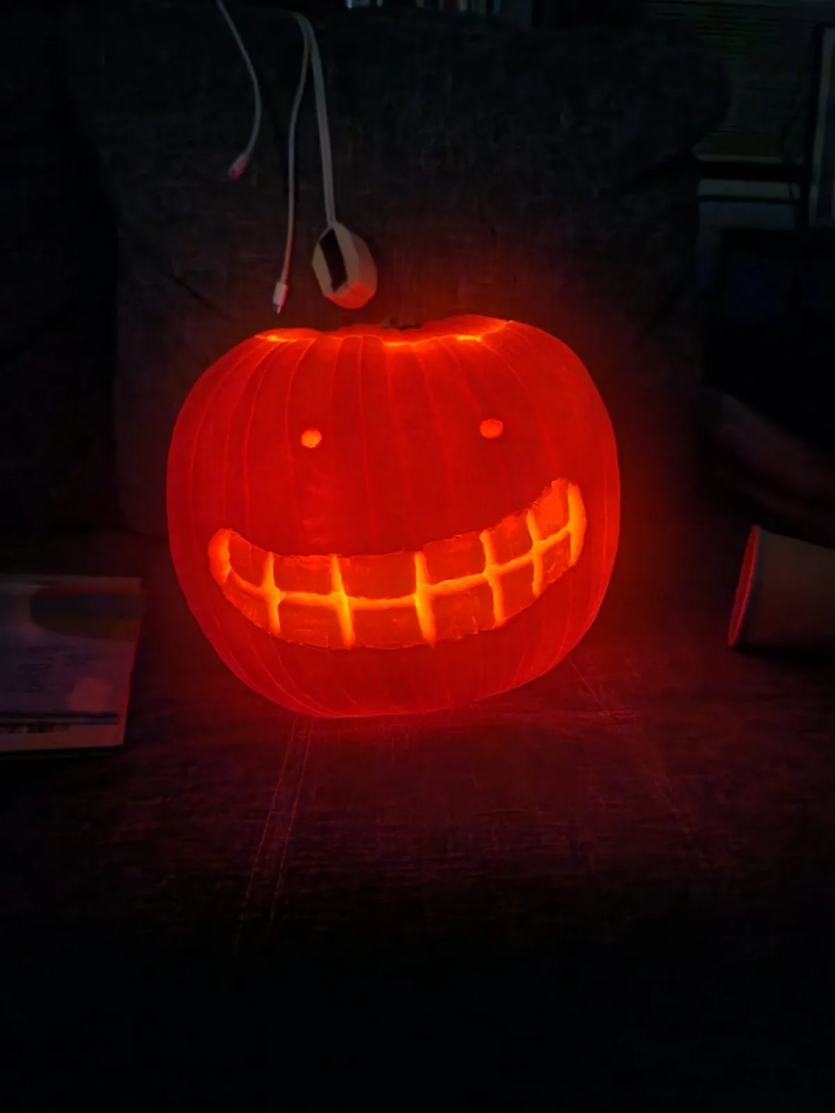 A carved pumpkin with a wide, toothy grin and two small eyes glows warmly from within, sitting on a dark surface. A USB…