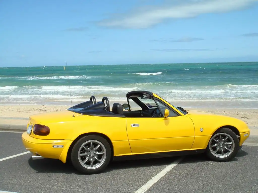 A bright yellow Mazda Miata convertible is parked in a lot near a beach with ocean waves visible in the background. The…