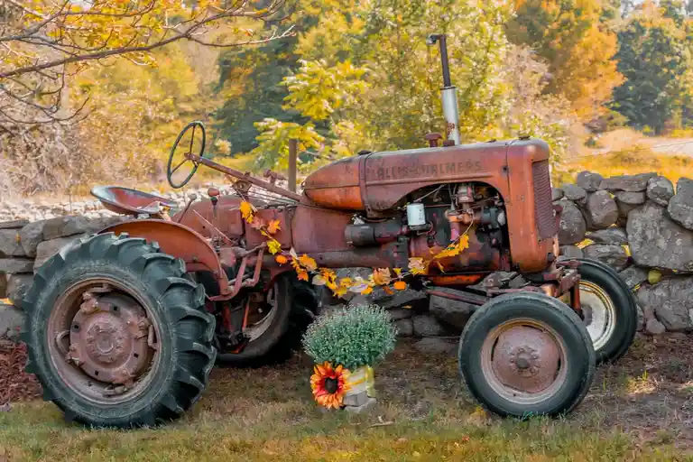 An old, rusty Allis-Chalmers tractor sits in a grassy field next to a stone wall, decorated with autumn leaves and a potted…