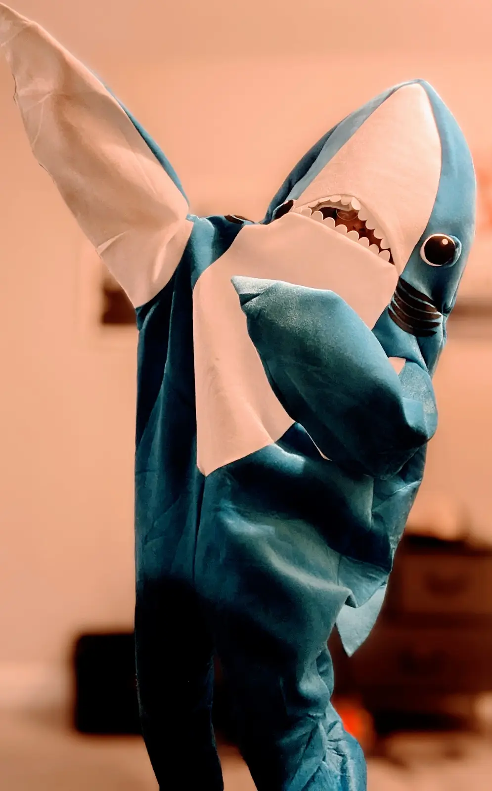 A person wearing a full-body shark costume with a large, toothy head and fin-like arms, standing with one arm raised. The…