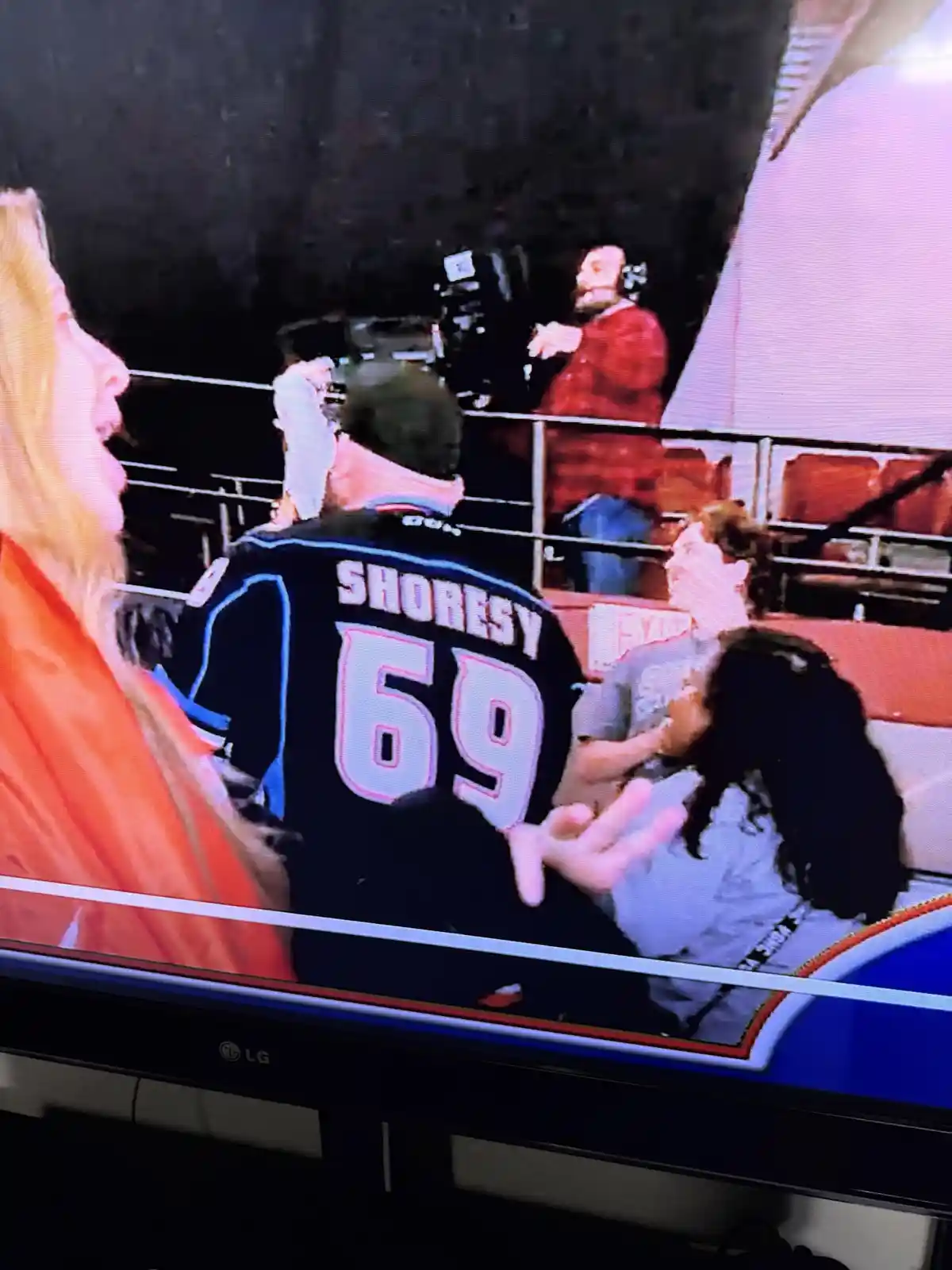 A television screen shows a wrestling event with a man in a black jersey with 'SHORESY 69' on the back, gesturing toward a…