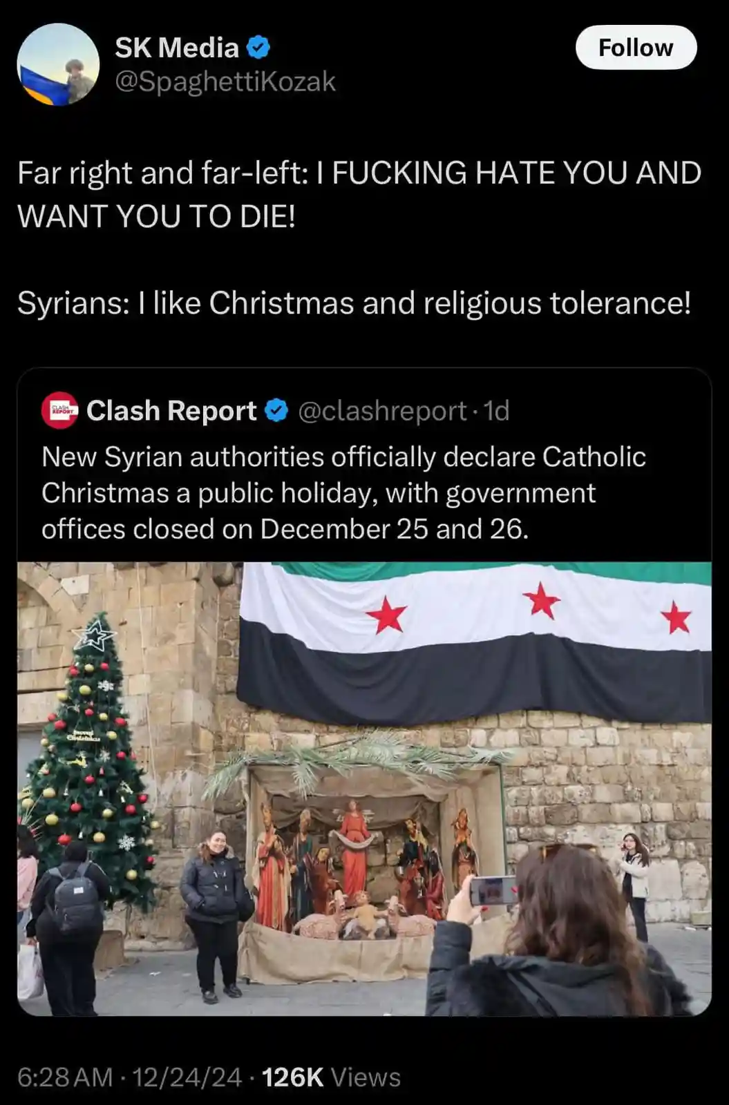 An image shows a public Christmas nativity scene with a decorated tree and a large Syrian flag hanging above it. People are…