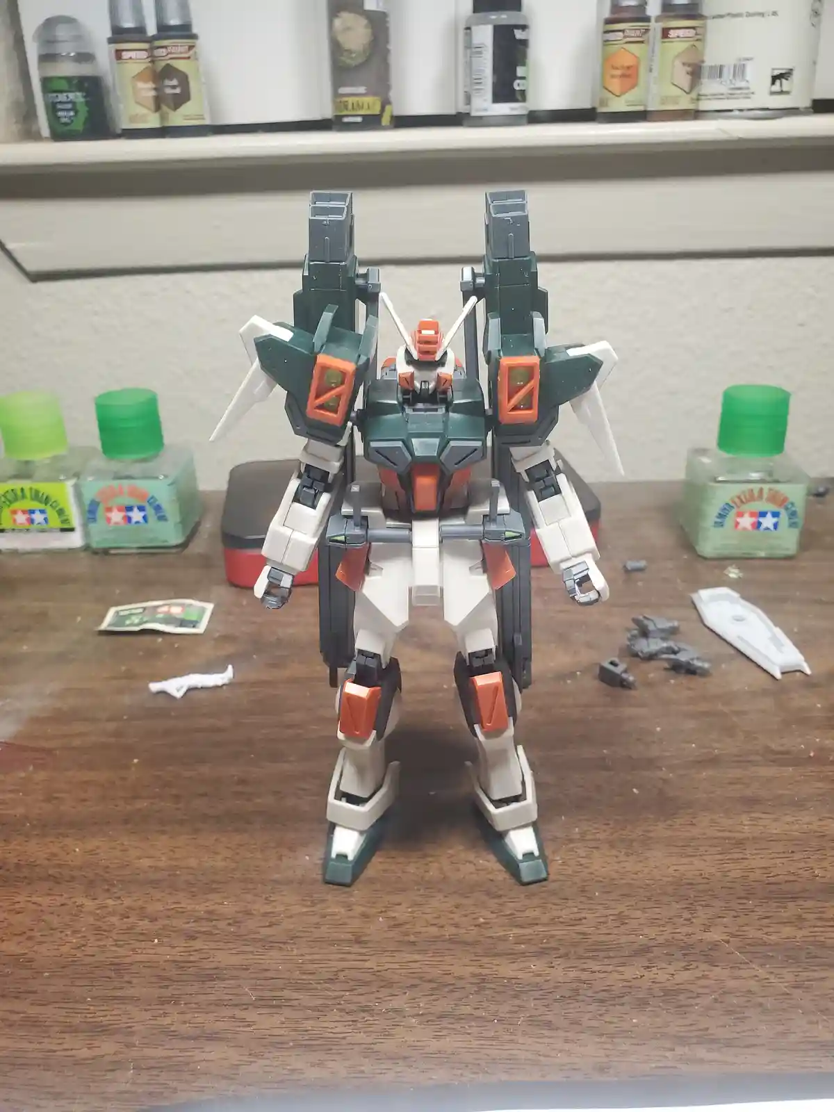 A green, white, and orange plastic model kit robot stands on a wooden workbench surrounded by model paints and parts. The…