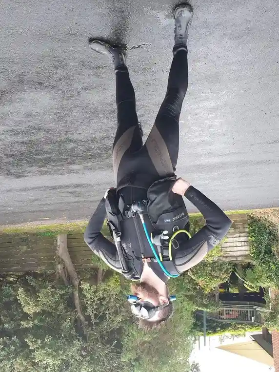 A man wearing full scuba gear, including a wetsuit, tank, and mask, is standing upright on a paved surface with his feet…