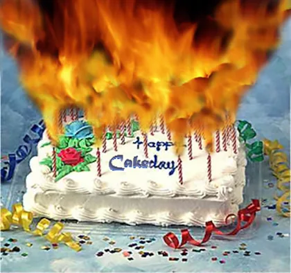 A rectangular white-frosted birthday cake with candles, a red rose decoration, and the text 'Happy Cakeday' is engulfed in…