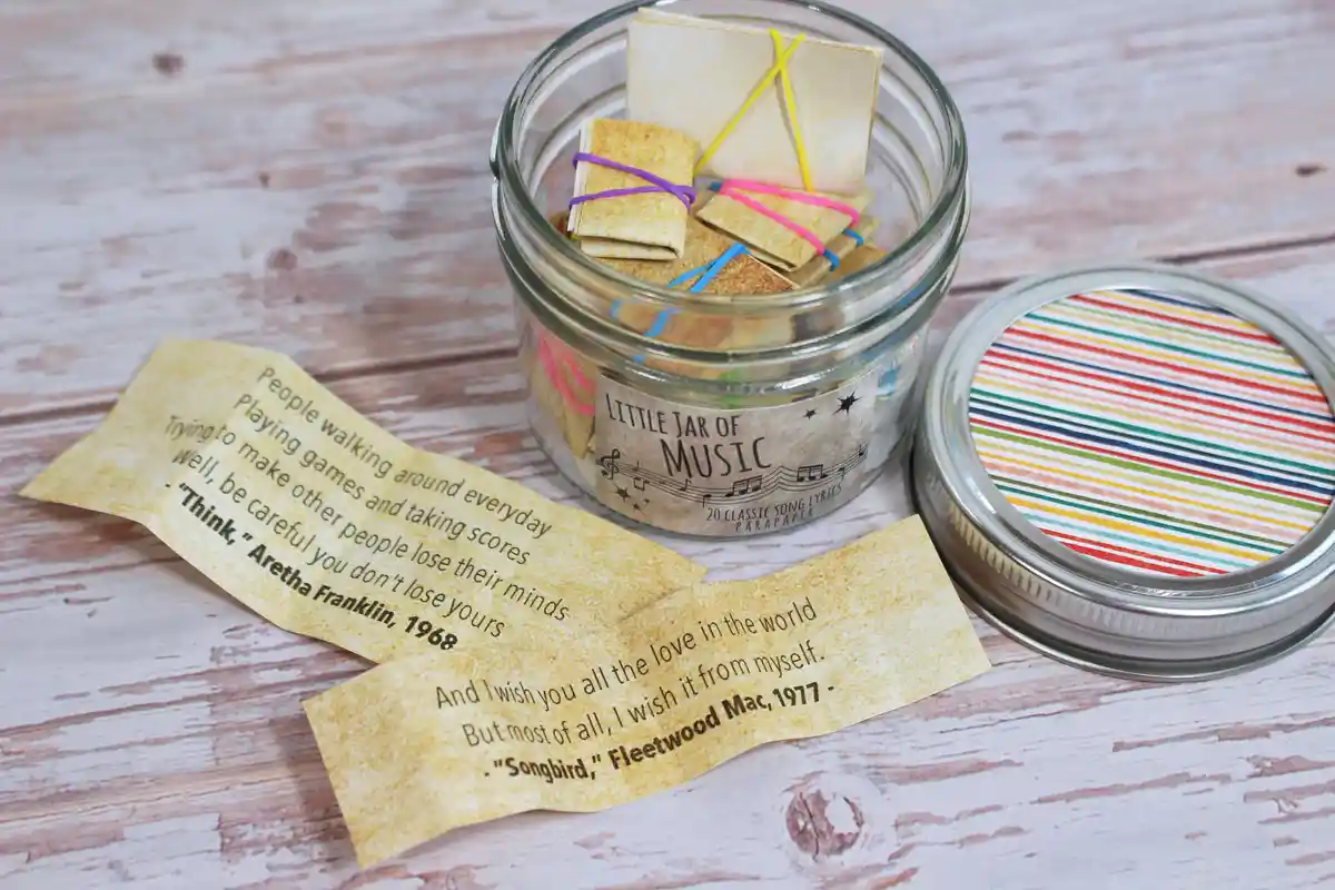 A glass jar labeled 'Little Jar of Music' contains small folded paper boats tied with colorful rubber bands, with two sample…