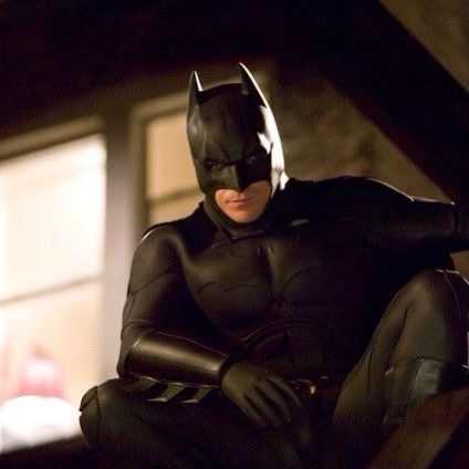 A man in a black Batman costume sits slumped in a dimly lit room, looking weary and defeated. The overlaid text reads 'i'm…