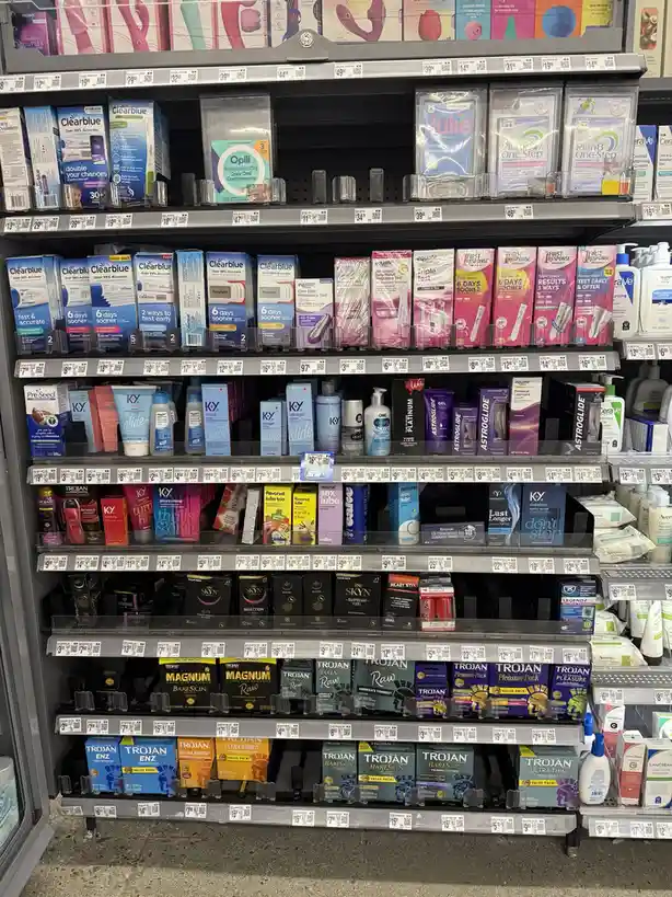 A photo of a well-stocked pharmacy aisle filled with pregnancy tests, condoms, and intimate care products. The top shelf…