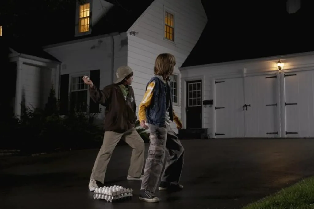Two young boys stand in a driveway at night, one holding a baseball bat while the other looks toward a house with lit…