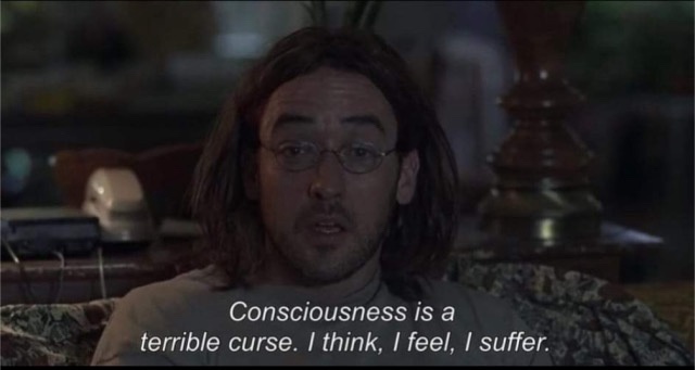A man with long hair and glasses sits on a couch in a dimly lit room, looking directly at the camera with a weary…