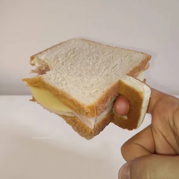 A hand holds a sandwich made with white bread, cheese, and deli meat, with a hot dog visibly protruding from the side. The…