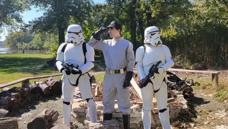 A person in a light gray uniform and black cap stands between two stormtrooper costumes, saluting with their right hand. The…