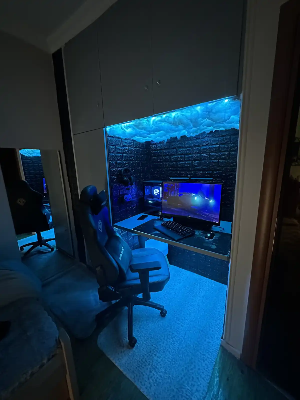 A dimly lit gaming room features a desk with a monitor, keyboard, and gaming chair, all bathed in blue light. The wall…