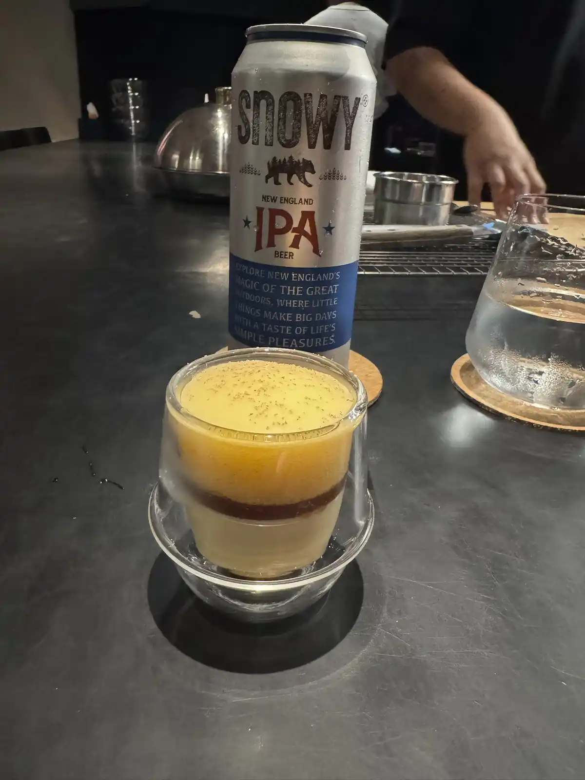 A chilled can of Snowy New England IPA beer stands behind a layered coffee drink in a glass on a dark countertop, with a…