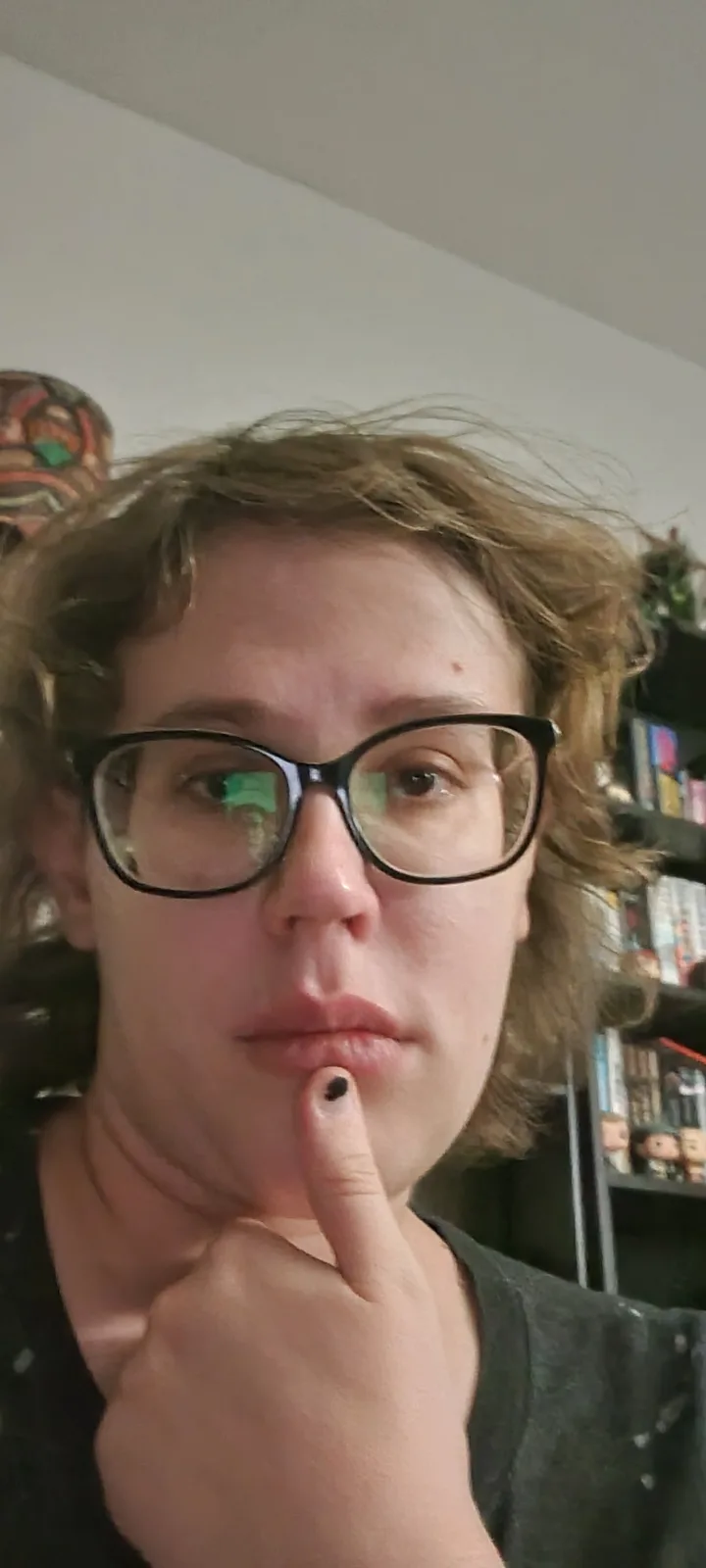 A person with curly brown hair and black-rimmed glasses holds their index finger vertically against their lower lip, looking…