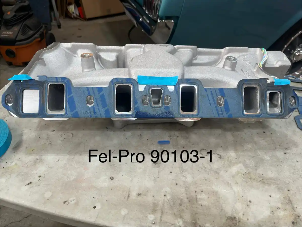 A silver aluminum automotive valve cover sits on a workbench, partially masked with blue painter's tape around its…