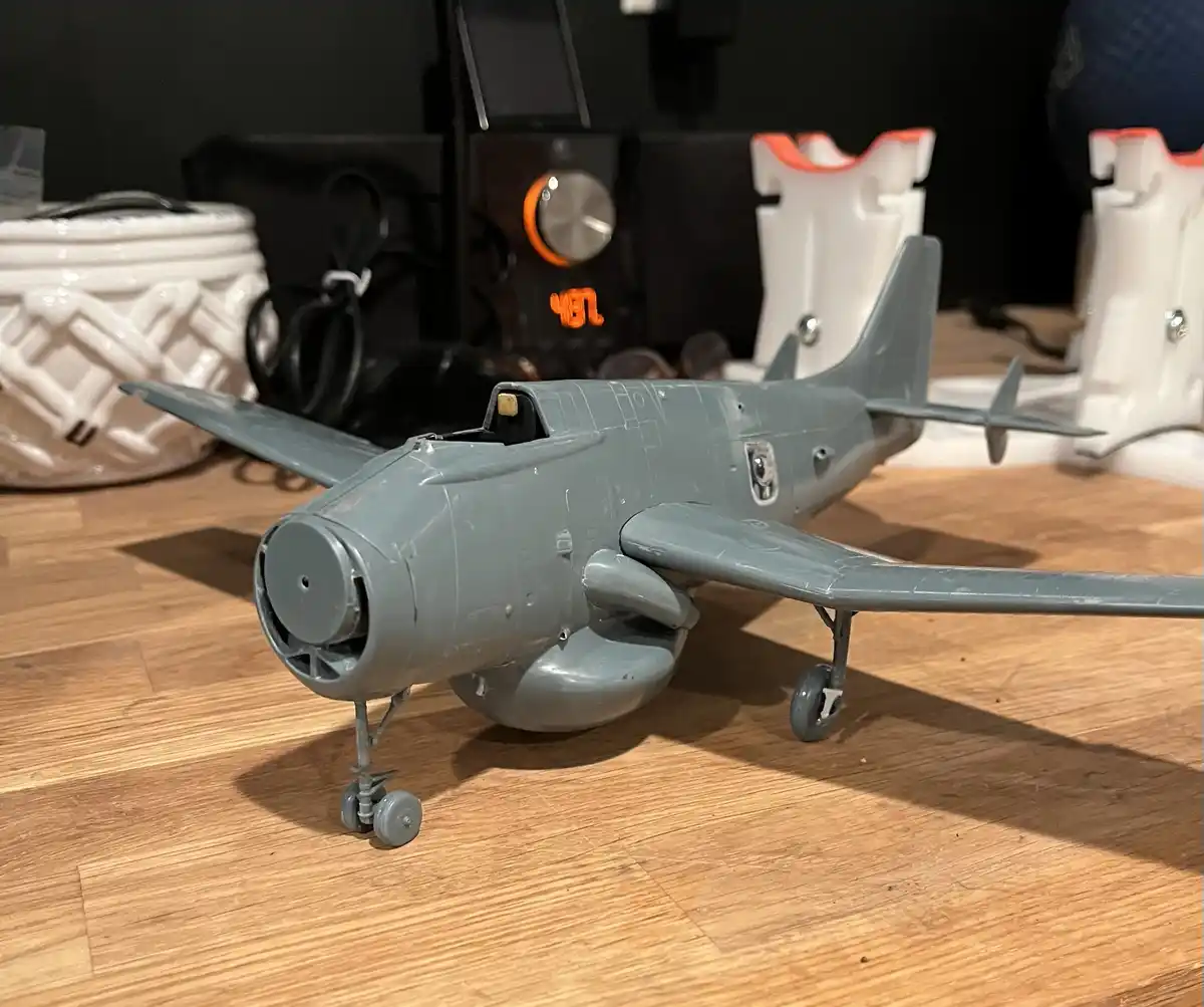 A grey, unpainted model airplane sits on a wooden floor, showing visible seams and unfinished details. In the background…