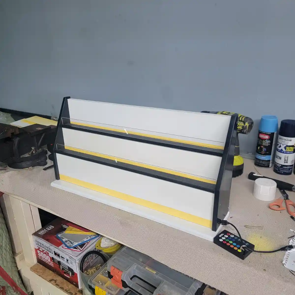 A white and black DIY shelf with yellow tape on its edges sits on a cluttered workbench surrounded by tools, spray paint…