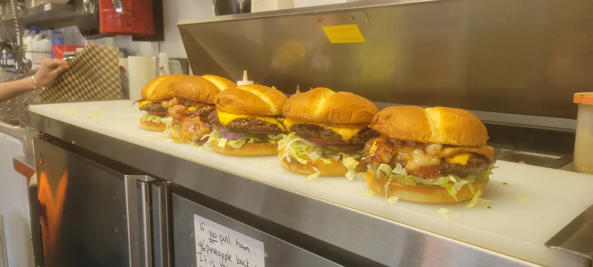 A row of five cheeseburgers with various toppings sits on a white cutting board in a commercial kitchen. The burgers vary in…