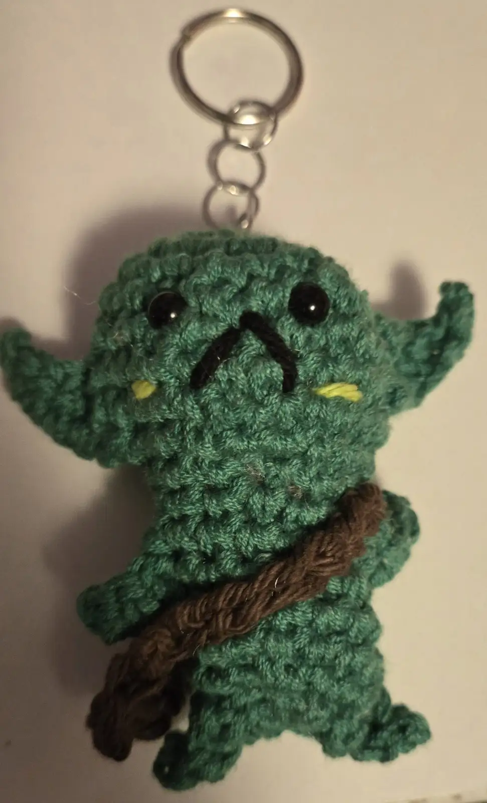 A handmade green crocheted keychain of the Star Wars character Yoda, featuring large black eyes, a stitched mouth, and a…