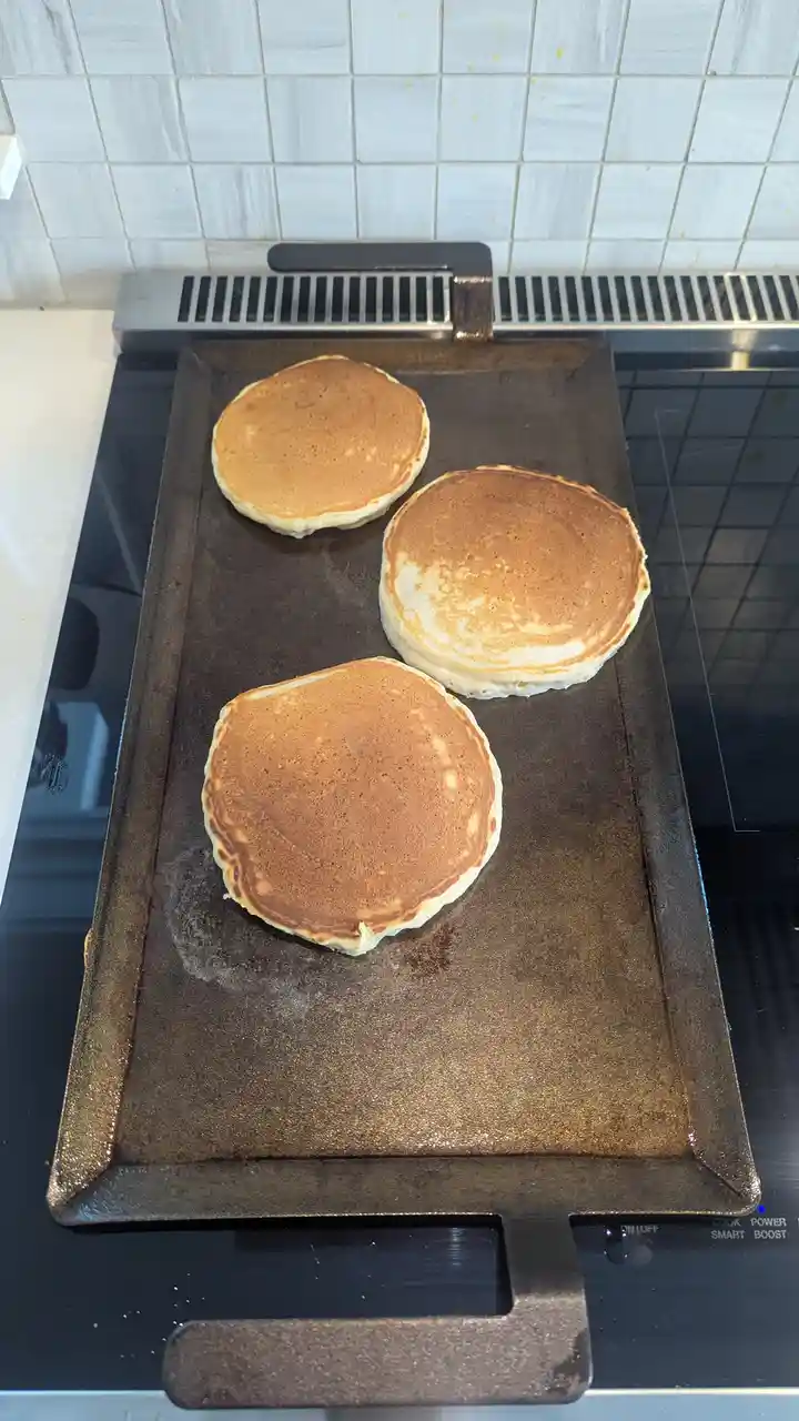 Three golden-brown pancakes are cooking on a well-used, dark metal griddle placed on a black glass stovetop, with a white…