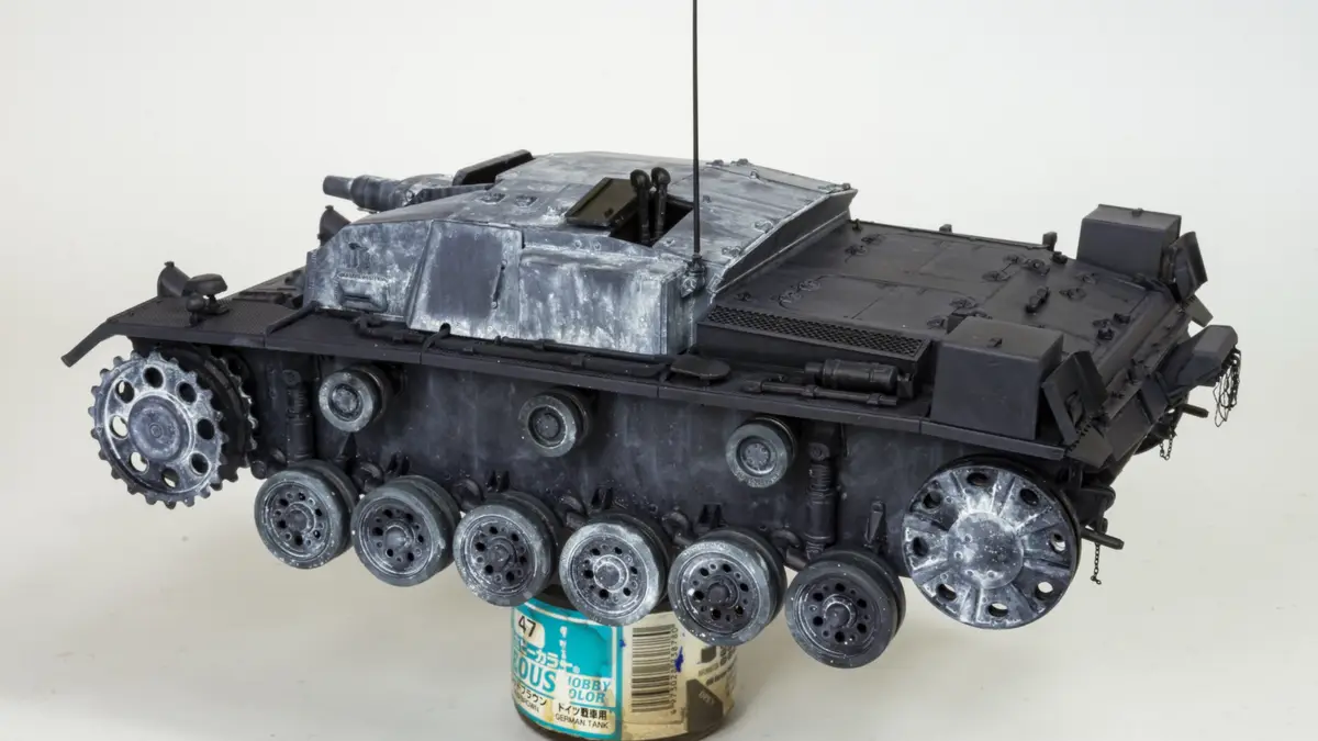 A detailed scale model of a German tank, likely a StuG III, is shown resting on a paint can labeled 'GERMAN TANK'. The tank…
