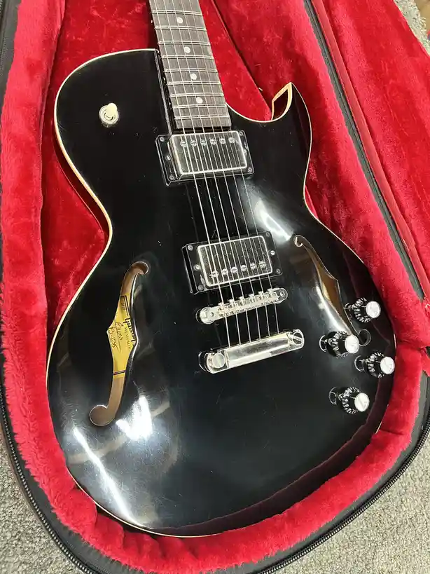 A black electric guitar with gold trim and two humbucker pickups rests inside a red velvet-lined case. The guitar has a…