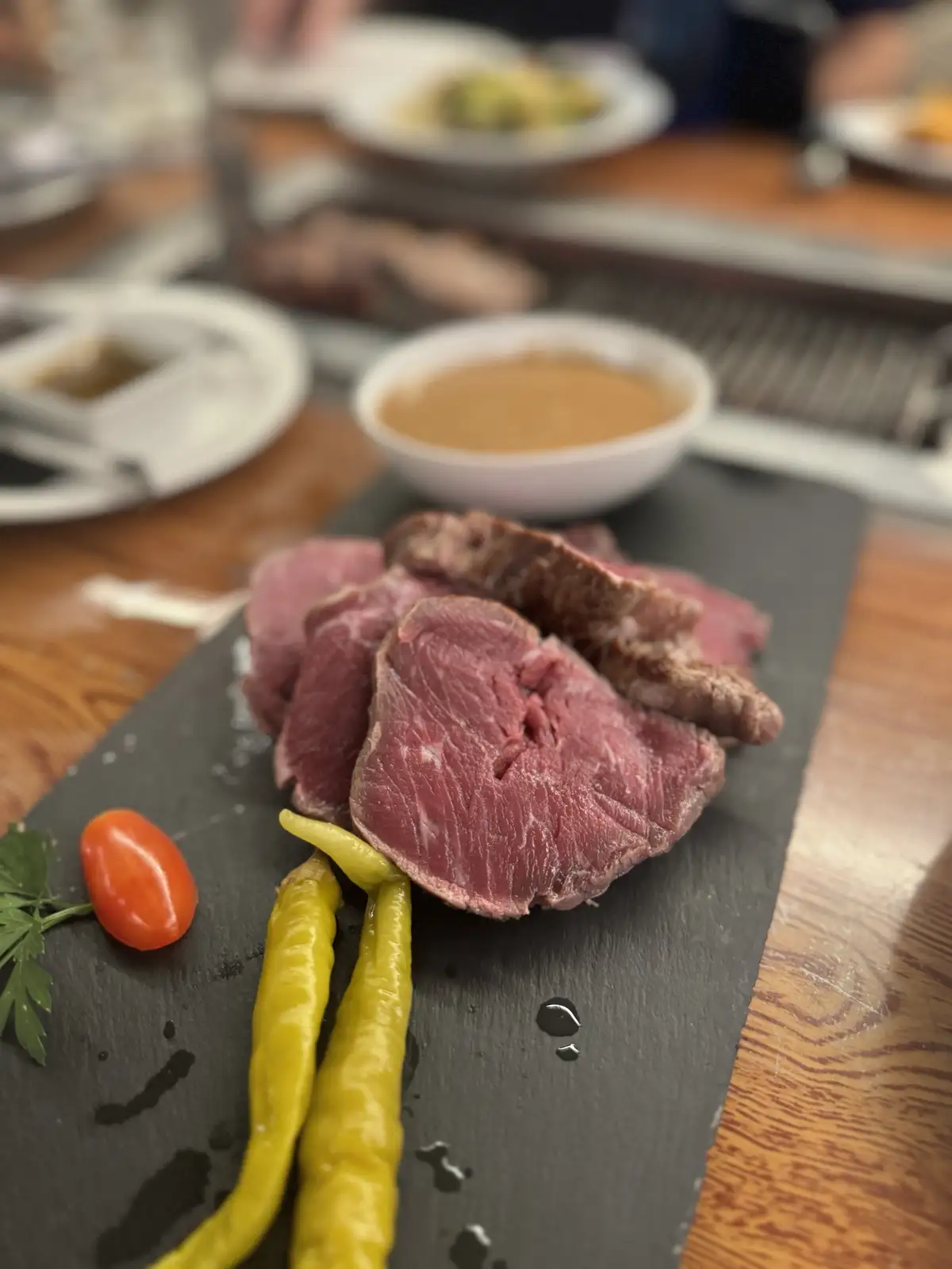 A close-up photo shows sliced medium-rare steak on a black slate plate, garnished with yellow peppers, a cherry tomato, and…