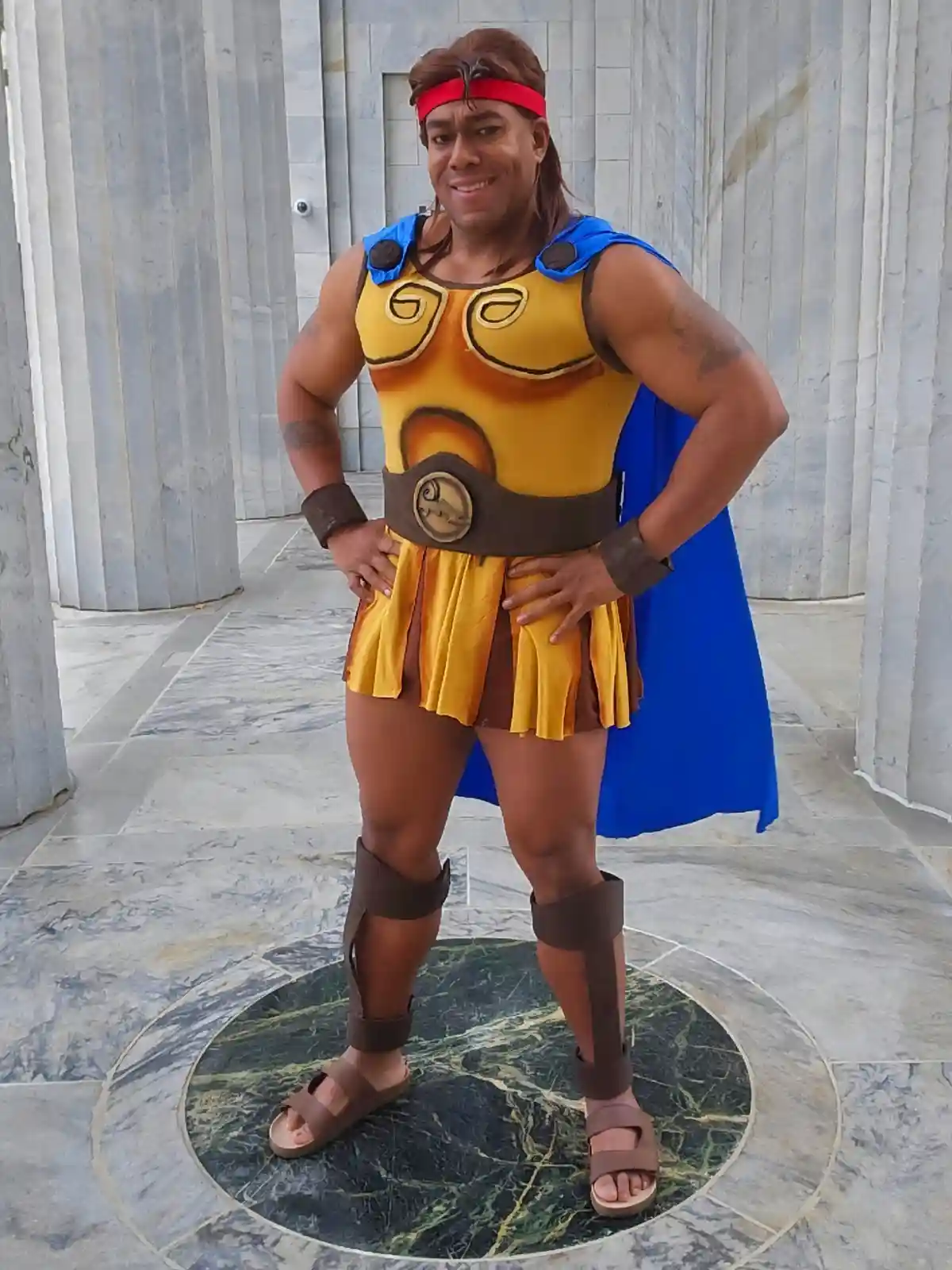 A muscular man in a yellow and blue Greek-style hero costume with a red headband, blue cape, and sandals poses confidently…