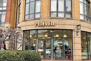 An exterior shot of a 7-Eleven convenience store with its iconic logo and name prominently displayed above the glass doors…