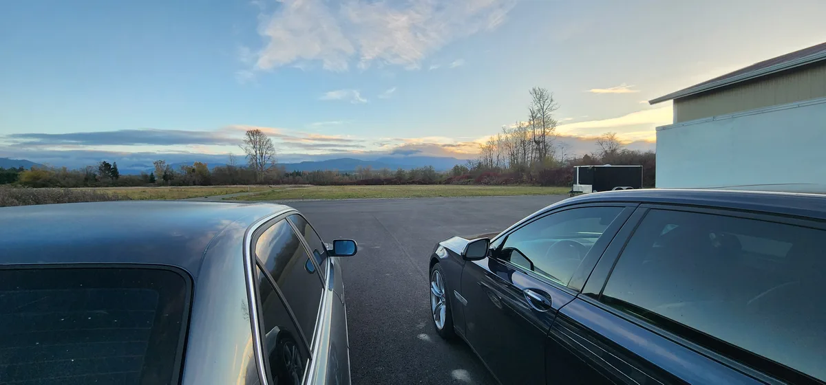 Two dark-colored cars are parked side-by-side in an open asphalt lot, facing a scenic view of distant mountains and a partly…