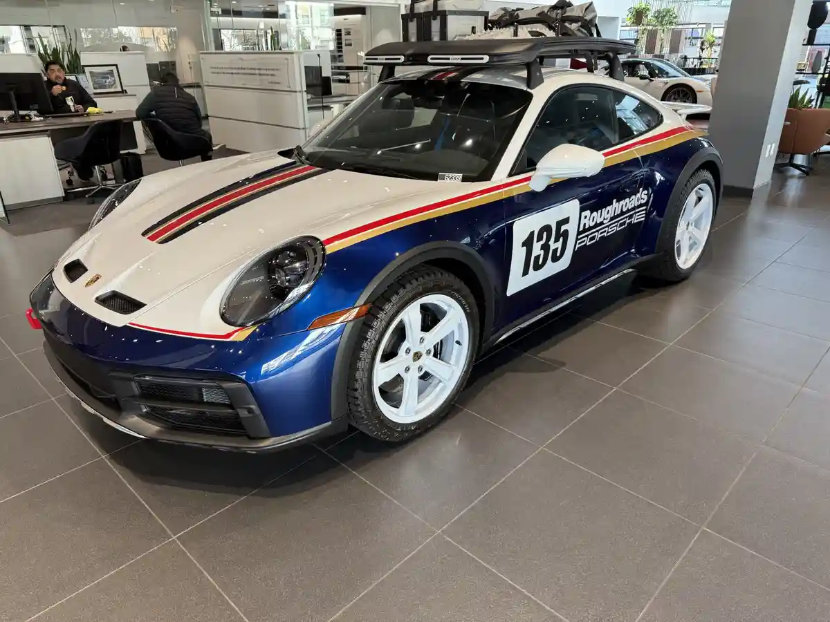 A blue and white Porsche 911 Dakar with '135 Roughroads PORSCHE' livery and off-road tires is displayed in a dealership…