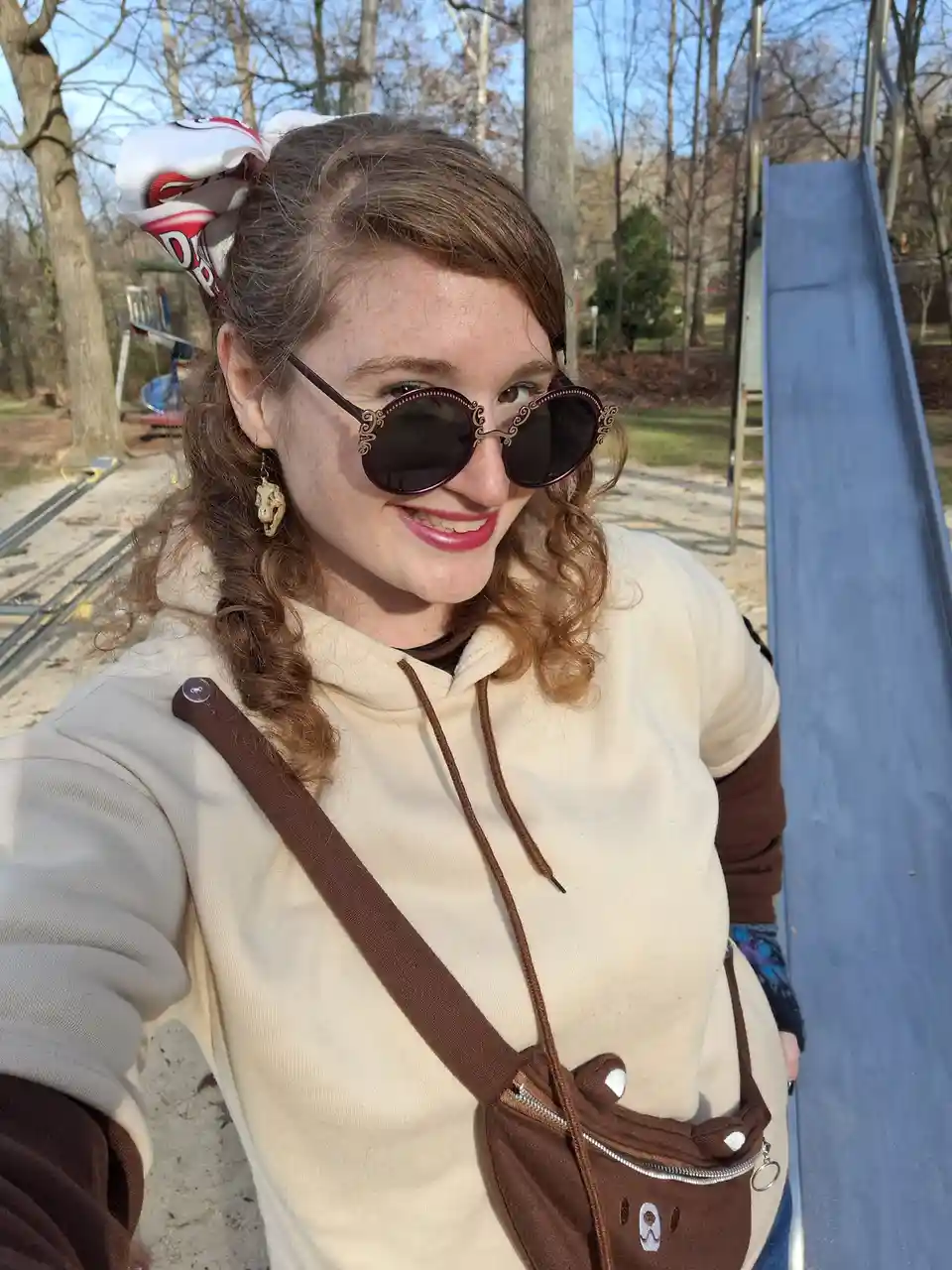 A young woman with curly brown hair, wearing ornate sunglasses and a beige hoodie, takes a selfie outdoors next to a blue…