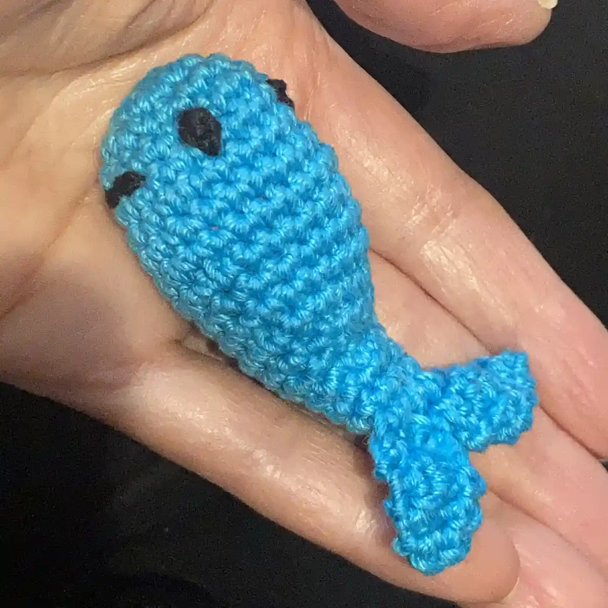 A small, blue crocheted fish with black eyes rests in the palm of a person's hand, showcasing the handmade craft. The…