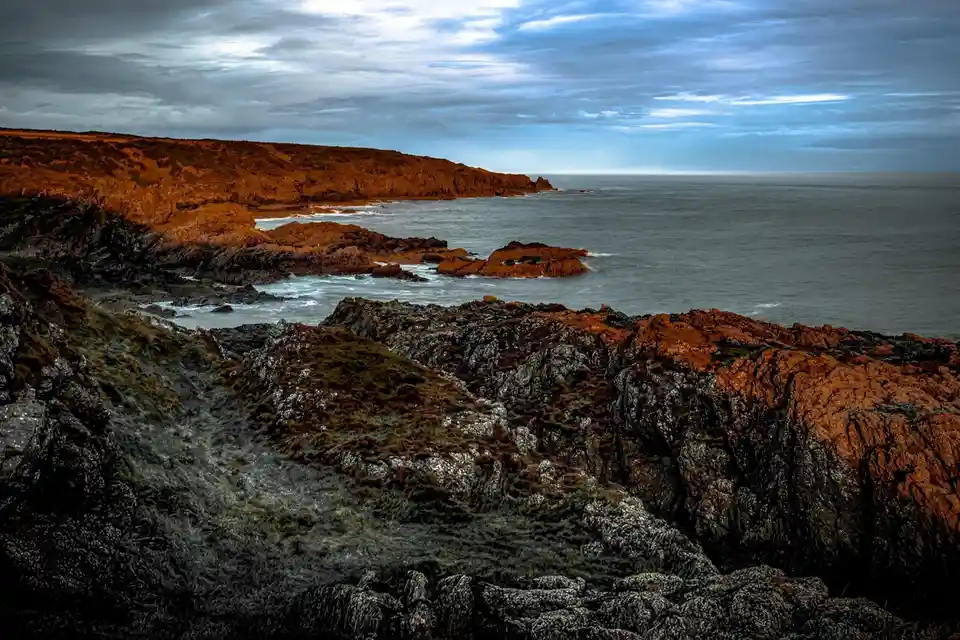 A dramatic coastal landscape with rugged, dark rocks in the foreground and a distant cliff line under a cloudy sky. The…
