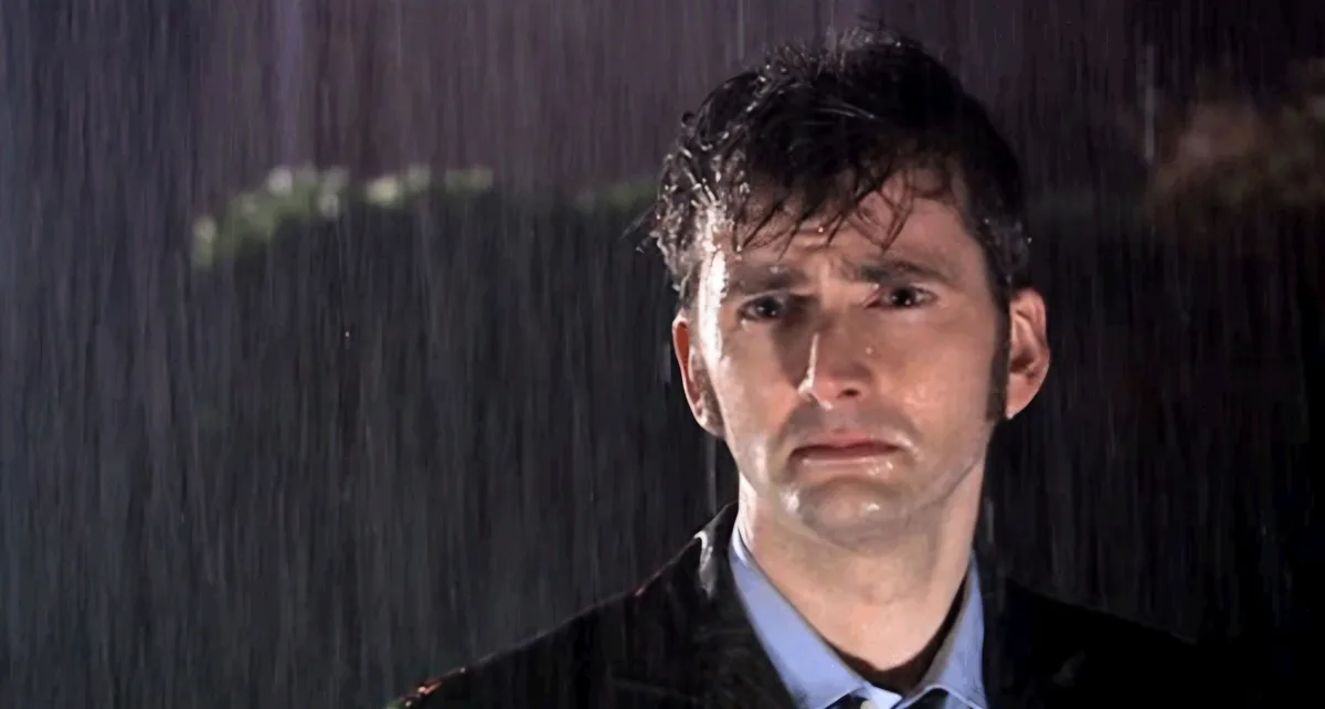 A man with wet, messy hair and a pained expression stands in heavy rain, water dripping down his face and coat. He wears a…