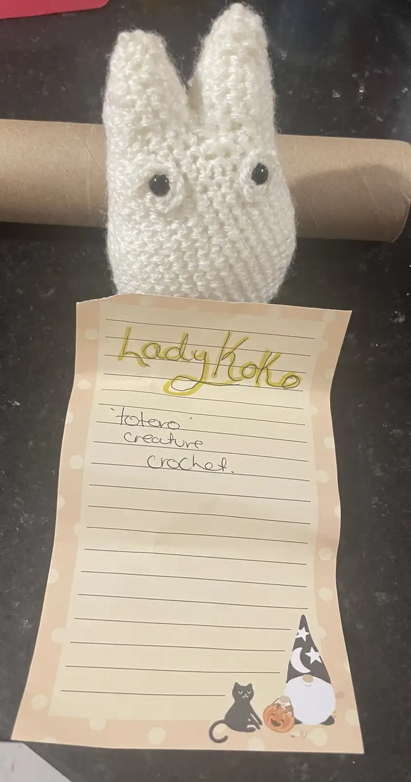 A white crocheted bunny head with black button eyes is peeking over a brown paper towel roll, holding a lined notepad. The…
