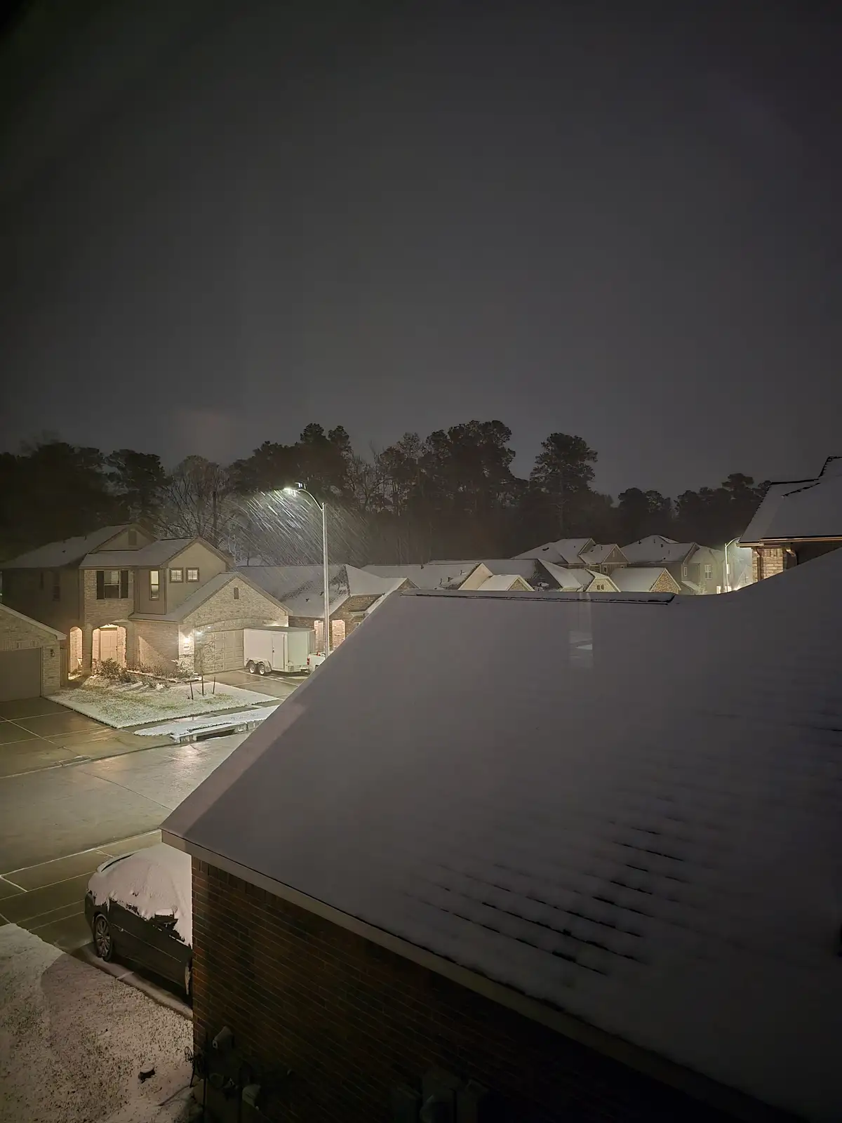 A nighttime photo of a suburban neighborhood blanketed in snow, with streetlights illuminating falling snowflakes and…
